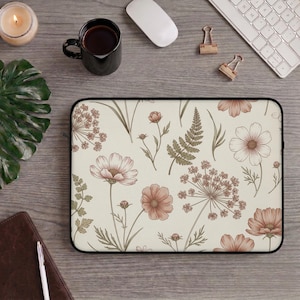 May include: A laptop sleeve featuring a cream-colored background with a floral pattern in shades of pink, brown, and green. The design includes detailed illustrations of flowers, leaves, and ferns. The sleeve has a black border and is designed to protect a laptop.
