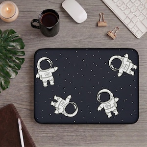 Puede incluir: Una funda rectangular para portátil con fondo azul marino oscuro, un patrón de estrellas blancas y cuatro ilustraciones de astronautas blancos. Cada traje de astronauta muestra una tecla de teclado: Alt, Esc, Ctrl y Space en negro.