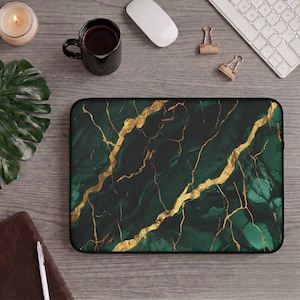 Könnte beinhalten: Eine Laptop-Hülle mit einem dunkelgrünen Marmormuster und goldenen Adern. Die rechteckige Hülle hat einen schwarzen Rand. Das Design ähnelt einer Geode oder einem Naturstein. Auf einem Holzschreibtisch mit Büroaccessoires.