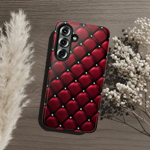 Op de afbeelding: Een smartphonehoesje met een dieprood, gewatteerd ruitpatroon met kleine, heldere, sprankelende accenten op elk kruispunt. Het hoesje heeft een verticale camera-uitsparing aan de linkerkant voor drie cameralenzen.