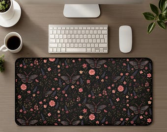 May include: A rectangular desk mat featuring a dark black background with a repeating pattern of dark grey moths, pink daisy-like flowers, small orange blossoms, and various green and purple botanical stems and leaves spread across the surface.