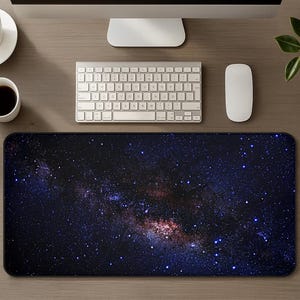 May include: A rectangular desk mat featuring a high-resolution print of the night sky, showcasing a dense field of stars, deep blue nebulae, and a prominent galactic core with shades of purple and orange against a dark black background.