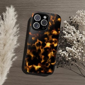 May include: A tortoiseshell-patterned phone case with a glossy finish. The case has a black border and a camera cutout for multiple lenses. The case is on a wooden surface with dried flowers and pampas grass.