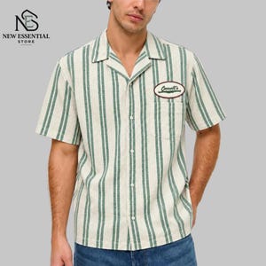 May include: A short-sleeve button-down shirt featuring a cream and green vertical striped pattern. The shirt has a notched collar, a single chest pocket, and an oval-shaped patch on the left chest with the text Cassell's in dark green.