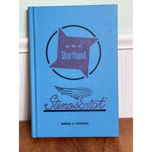 May include: A light blue hardcover book titled Stenoscript by Manuel C. Avancena. The cover features a dark purple four-pointed star shape containing the text a-b-c Shorthand, a circular outline, and a stylized wing graphic above the title.