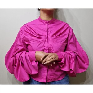 Op de afbeelding: Een felroze blouse met een opstaande kraag en lange, volumineuze mouwen. De blouse heeft een geplooid ontwerp over de borst en een knoopsluiting aan de voorkant. De mouwen zijn bij de manchetten geplooid, wat een dramatische, oversized look creëert. De blouse is gemaakt van een gladde stof.