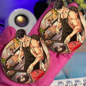 May include: Two circular enamel pins featuring an anime-style illustration of a person with dark hair wearing a black apron while cooking in a kitchen. The scene includes a red pot, a frying pan with an egg, and a wooden cutting board.