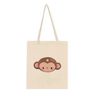 Peut inclure: Un sac fourre-tout en toile beige avec une illustration graphique centrée représentant une tête de singe marron ornée d'une petite étoile jaune sur le front. Le singe a de grandes oreilles rondes, des joues roses et un visage simple.