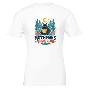 Può includere: Una t-shirt bianca a maniche corte con la grafica di una creatura blu scuro simile a una falena che legge un libro giallo. Il disegno include pini, una luna crescente, stelle e il testo "MOTHMAN'S BOOK CLUB" in caratteri blu e arancioni.