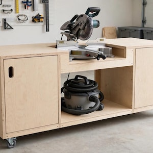 Puede incluir: Un banco de trabajo de madera contrachapada de color claro sobre ruedas con dos armarios laterales con tiradores recortados, una plataforma central empotrada con una sierra ingletadora y un estante inferior abierto con una aspiradora.
