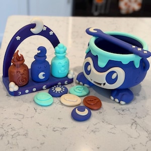 May include: A dark blue monster-shaped cauldron with a light blue rim and spoon, accompanied by a purple arch stand holding three small potion bottles in brown, blue, and light blue, plus several circular tokens with various embossed symbols.