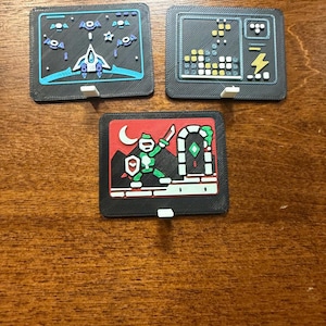 May include: Three rectangular black plastic wall mounts featuring raised, multi-colored 3D printed designs. One shows a space ship, another a block puzzle game, and the third a green knight character standing before a stone archway.