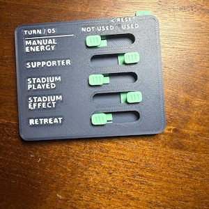 May include: A dark gray rectangular game tracker with light green sliding tabs. The board features text labels including TURN / 05, MANUAL ENERGY, SUPPORTER, STADIUM PLAYED, STADIUM EFFECT, RETREAT, NOT USED, USED, and RESET. The surface has a textured finish.
