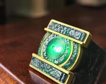 Può includere: Un anello caratterizzato da un elemento luminoso circolare verde centrale circondato da una cornice color oro. La fascia è grigio scuro con una superficie ruvida e usurata. Il design include pannelli rettangolari sopra e sotto il centro verde.
