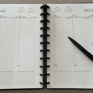 May include: An open black disc-bound planner with white pages featuring a weekly layout dated DEC 28 - JAN 03. The planner includes hourly slots, sections for priorities, and a black ballpoint pen resting diagonally across the right page.