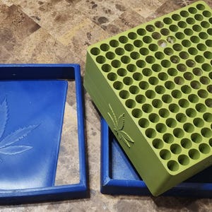 May include: A blue square tray with a debossed cannabis leaf design on the base and a separate lime green square block featuring a grid of one hundred circular holes, with a matching cannabis leaf emblem embossed on the side of the green block.