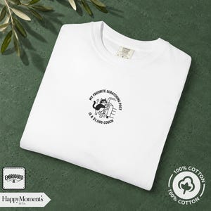 Könnte beinhalten: Ein weißes T-Shirt mit einer kleinen schwarzen Stickerei in der Mitte. Das Motiv zeigt eine Katze, die an einem Sofa kratzt, mit dem Text "MY FAVORITE SCRATCHING POST IS A $1,500 COUCH", der kreisförmig um die Illustration angeordnet ist.