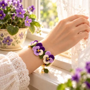 May include: A crochet bracelet featuring multiple pansy flower motifs in shades of purple, lavender, and white with yellow centers, accented by small green leaves, a gold-toned chain, and a single white pearl bead charm.