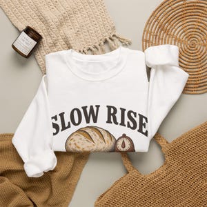 Puede incluir: Una sudadera blanca de manga larga con cuello redondo que presenta el texto "SLOW RISE" en letras mayúsculas de color marrón oscuro. Debajo del texto hay una ilustración gráfica de una hogaza de pan redonda y un temporizador de cocina marrón.