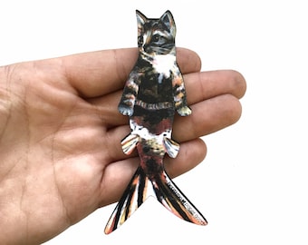 Puede incluir: Una ilustración colorida de una sirena gato con una cabeza de gato negra, marrón y blanca y una cola de pez colorida. La ilustración está impresa en un marcapáginas de madera con el texto "handmade paper goods" en la parte inferior.