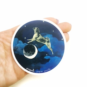 May include: A round sticker with a white border features a pug dog jumping in front of a crescent moon and stars on a blue and purple night sky. The text "pergamino paper goods" is printed in white at the bottom of the sticker.