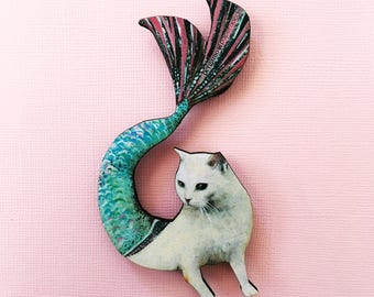 May include: A white cat with a teal and pink mermaid tail. The cat is sitting on its hind legs and looking to the left. The tail is curled around the cat's body.
