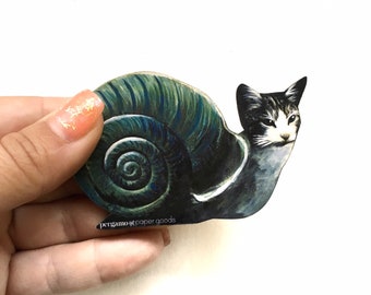May include: A black and white cat with green eyes is depicted as a snail. The snail shell is a swirling pattern of dark blue and green. The image is a sticker with the text "pergamost paper goods" at the bottom.