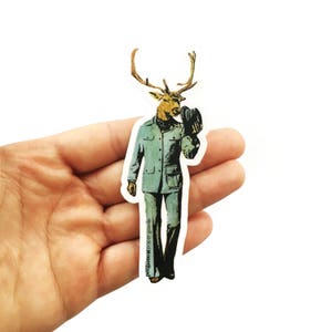 May include: A sticker featuring a deer with a human body wearing a blue jacket and pants. The deer is holding a black hat in its hand. The sticker has the text "pretty wild paper goods" at the bottom.