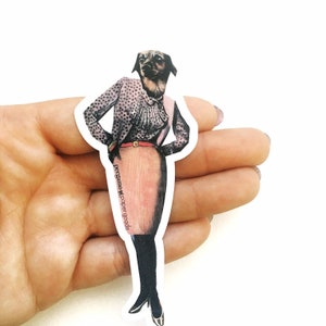 Puede incluir: Una pegatina de un perro con una blusa de lunares rosa y negro, un cinturón rojo y una falda rosa. El perro lleva tacones negros y tiene una expresión seria. La pegatina está sobre un fondo blanco y tiene el texto "pergamino paper goods" impreso en la parte inferior.