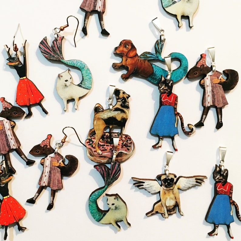 May include: Assortment of colorful wooden earrings and pendants featuring whimsical animal and mermaid designs. The earrings and pendants are made of wood and feature hand-painted details. The designs include a cat, a dog, a mermaid, a pug, and a squirrel.