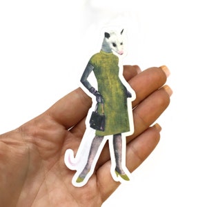 May include: A sticker of a possum wearing a green dress and holding a black purse. The possum has a white face and a pink tail.