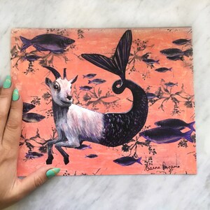 Puede incluir: Una ilustraci&oacute;n caprichosa de una cabra con cola de pez, sobre un fondo de color coral con un patr&oacute;n de peces morados. La firma del artista, "Barna Bagano", est&aacute; en la esquina inferior derecha.