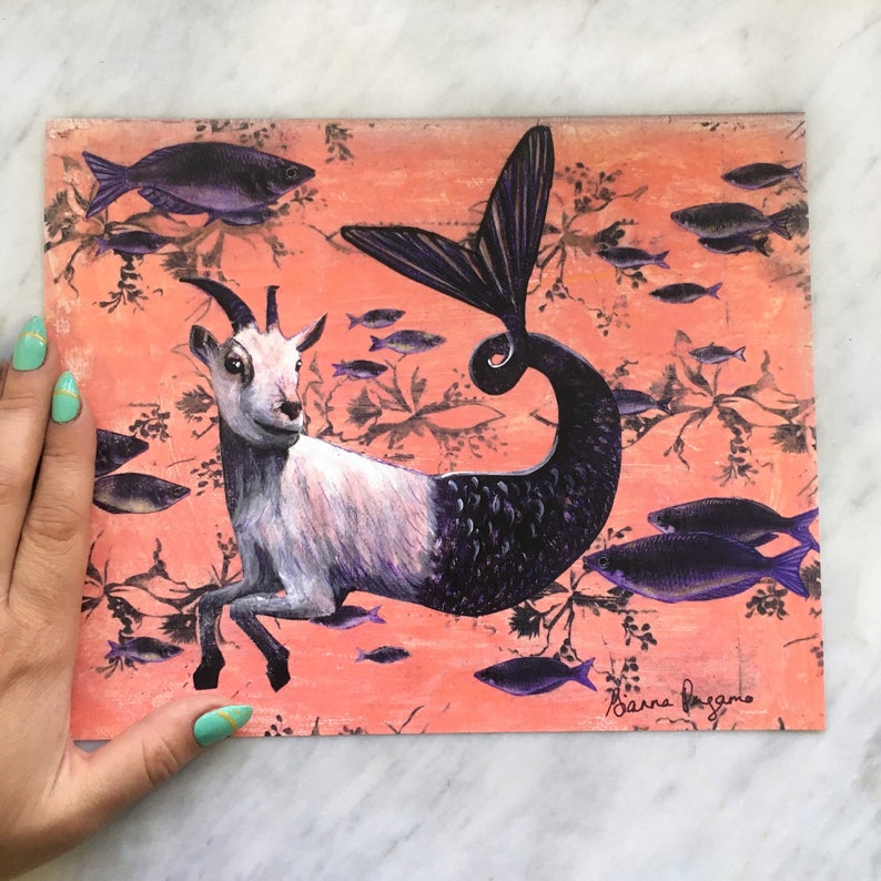 Puede incluir: Una ilustraci&oacute;n caprichosa de una cabra con cola de pez, sobre un fondo de color coral con un patr&oacute;n de peces morados. La firma del artista, "Barna Bagano", est&aacute; en la esquina inferior derecha.