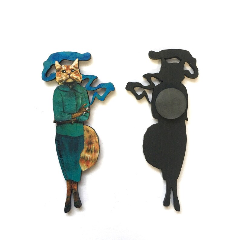 May include: A wooden magnet featuring a cat with a blue scarf and a cigarette, with a black silhouette of the same design on the back.