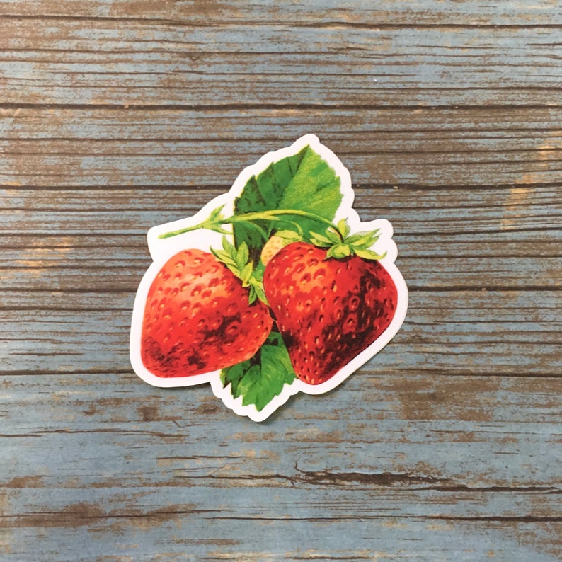 May include: A sticker depicting two red strawberries with green leaves and stems. The strawberries have textured details, and the leaves are a vibrant green. The sticker is on a blue and brown wooden surface.