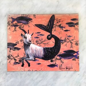 Puede incluir: Una ilustraci&oacute;n caprichosa de una cabra con cola de pez, rodeada de peces morados sobre un fondo coral. La obra est&aacute; firmada "Anna Fragos".
