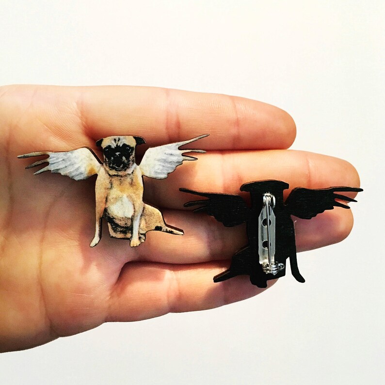 May include: A wooden pin featuring a pug with angel wings. The back of the pin shows a black silhouette of a bird with wings spread.
