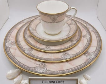 May include: A fine bone china set featuring a pink marble pattern with shell motifs and gold trim. The set includes a teacup, saucer, and three stacked plates of varying sizes. A sign reads: FINE BONE CHINA RARE PINK MARBLE SHELL DESIGN.