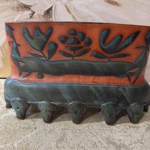 May include: A rectangular ceramic planter with a burnt orange upper section featuring dark blue incised floral and bird motifs. The lower base is a dark, mottled blue-green glaze with a series of six protruding, sculpted feet along the bottom.
