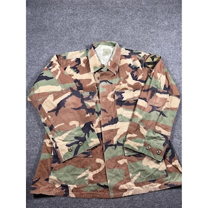 May include: A long-sleeve button-up shirt featuring a woodland camouflage pattern in shades of brown, green, and black. The shirt includes a pointed collar, two chest pockets with buttoned flaps, and adjustable buttoned cuffs on the sleeves.