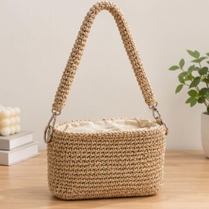 May include: A beige rectangular crochet handbag featuring a thick, textured woven handle attached with silver-toned metal rings. The bag has a soft, light-colored fabric interior lining visible at the top opening, showcasing a structured, hand-crafted design.
