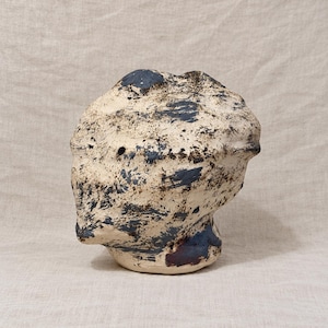 May include: A sculptural ceramic head with an irregular, textured surface. The piece features a cream-colored base glaze heavily mottled with dark blue and black splatters, creating a rugged, weathered appearance with a small circular opening.