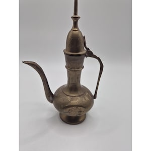 May include: A metallic brass oil pourer featuring a rounded base, a slender neck, and a long curved spout. The surface is decorated with etched floral patterns and includes a tall, pointed lid with a hinged handle attached to the side.