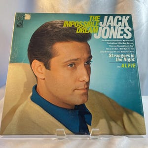 May include: Vintage vinyl record album cover featuring Jack Jones. The album is titled "The Impossible Dream" and includes song titles like "Strangers in the Night" and "Alfie". The cover art features a portrait of Jack Jones in a blue shirt and a cream-colored sweater.