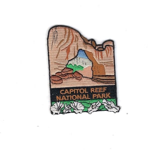 May include: An embroidered patch featuring a landscape of Capitol Reef National Park with tan rock formations, a blue sky, and green foliage. The text CAPITOL REEF NATIONAL PARK is stitched in orange on a black background above white flowers.
