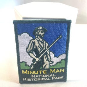 May include: Embroidered patch featuring a colonial-era figure with a rifle, set against a blue sky and green landscape. The patch is bordered in dark green and includes the text "MINUTE MAN NATIONAL HISTORICAL PARK" in yellow and white.