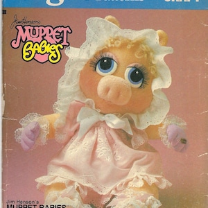 May include: A vintage Vogue Patterns craft sewing pattern featuring a pink pig Muppet doll wearing a white lace dress and bonnet. The pattern is titled "Jim Henson's Muppet Babies Soft Sculpture & Clothes" and includes the text "8967 CRAFT".