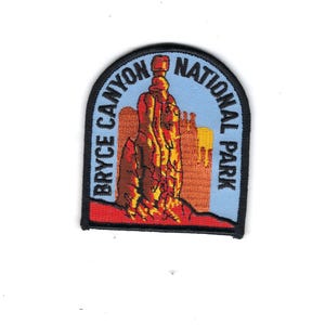 May include: An embroidered patch featuring a light blue sky background with a central red and orange rock formation. The text BRYCE CANYON NATIONAL PARK is stitched in black along the curved border. The patch has a thick black stitched edge.