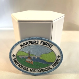May include: An oval patch featuring the text "HARPERS FERRY NATIONAL HISTORICAL PARK" in white lettering. The patch depicts a river scene with a bridge, buildings, and green hills, all within a blue border.
