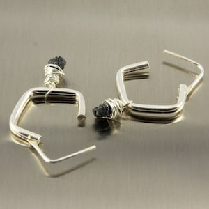 May include: A pair of silver-coloured square hoop earrings. Each earring has a small, dark, rough-textured stone wrapped in silver wire at the top. The hoops have a simple, modern design with a post closure.